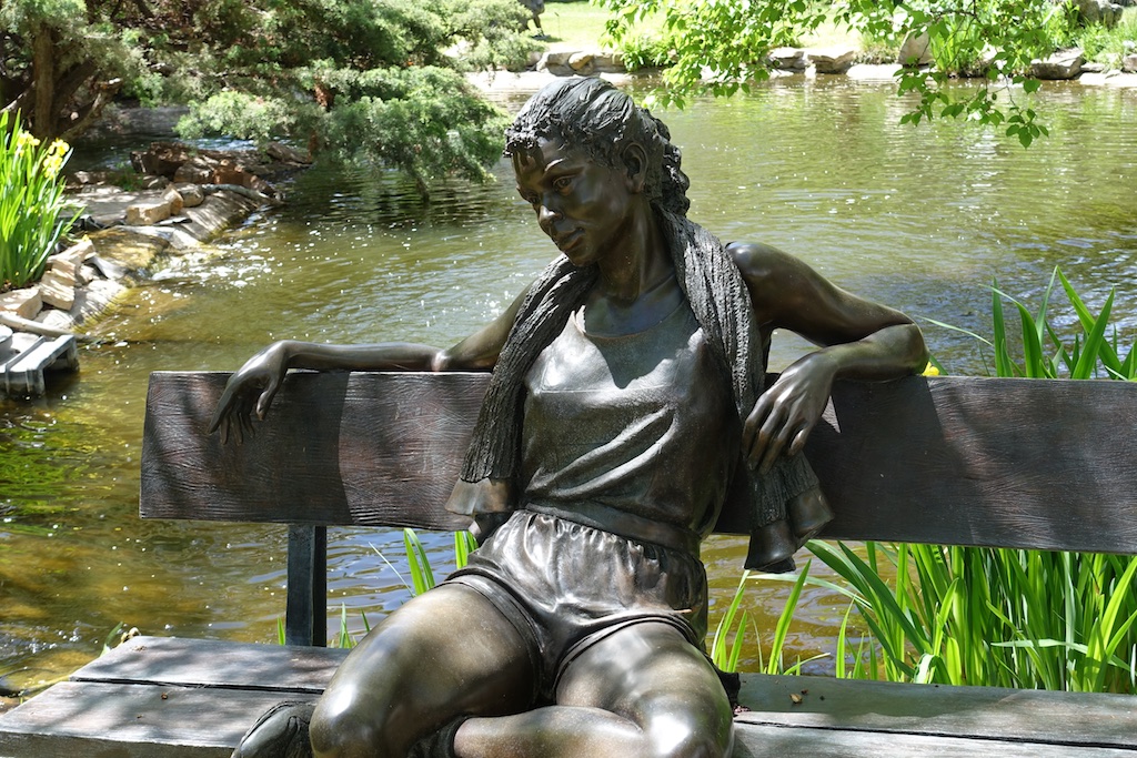 Sculpture of a woman sitting on a park bench with a pond behind.