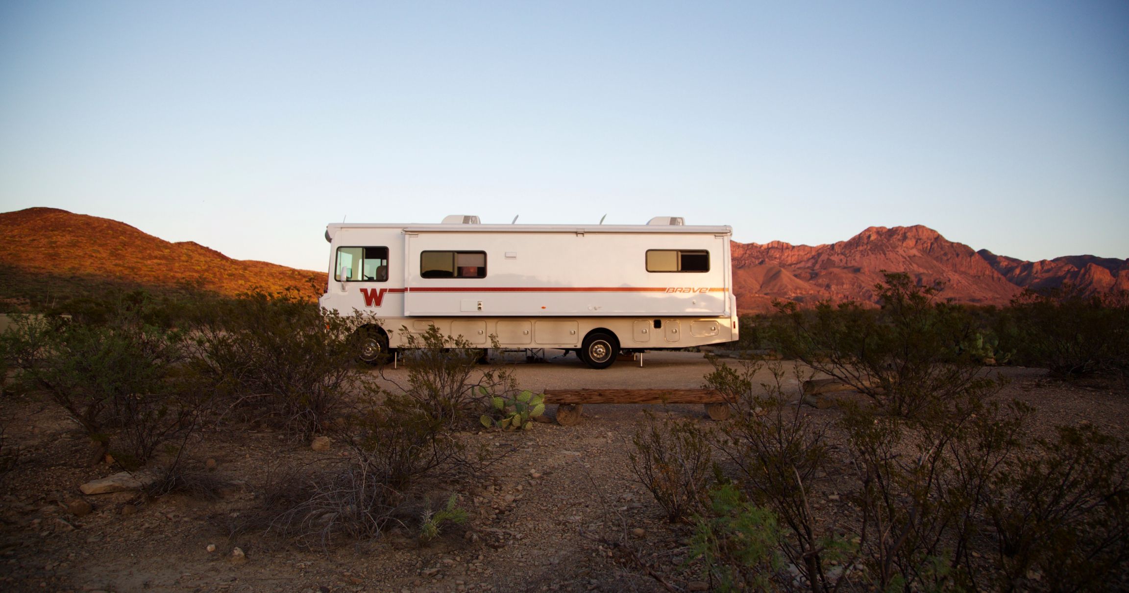Winnebago Brave parked in a camp site surrounded by desert terrain and red rocky mountains in the background.