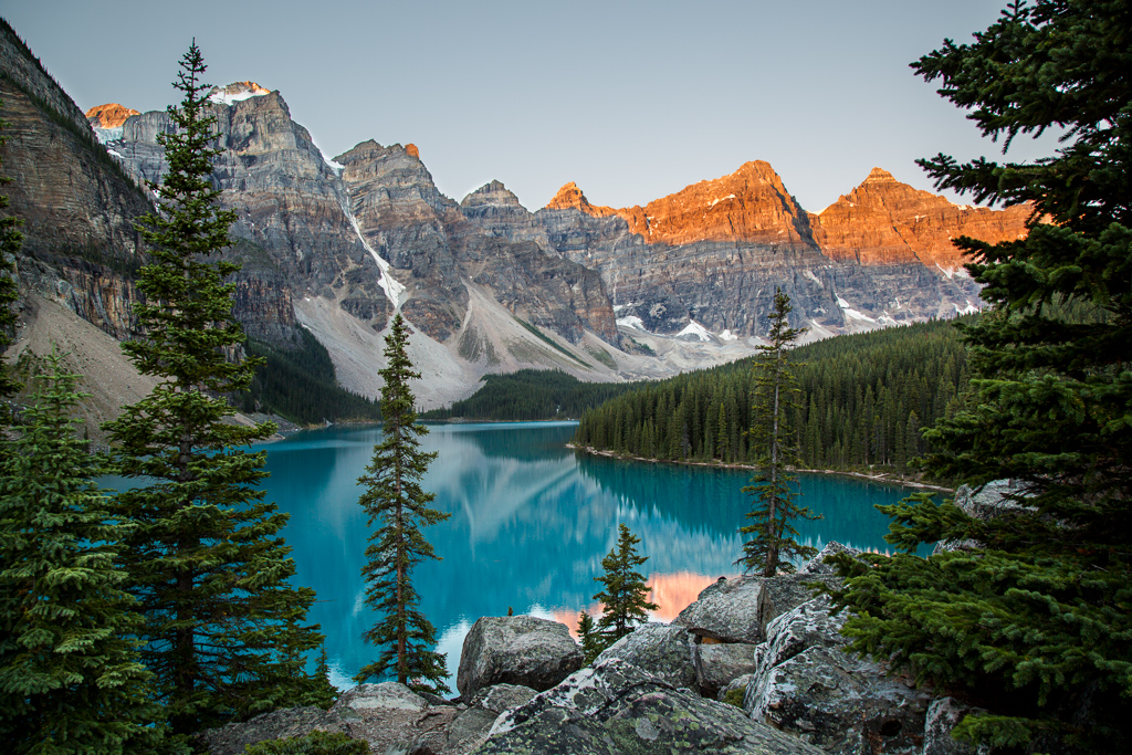 Clear blue Moraine Lake surrounded by trees with sun just hitting the tops of the mountains. 