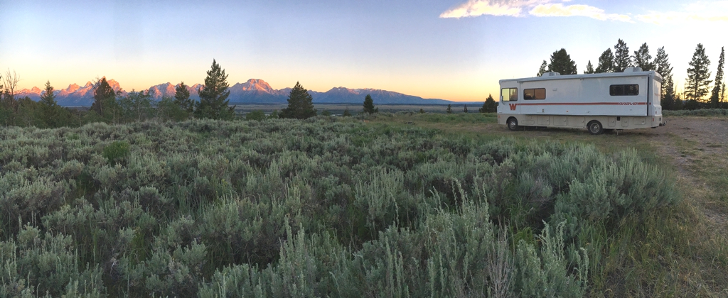 Winnebago Brave parked in a grassy site with the sun hitting the tops of the Tetons in the distance.