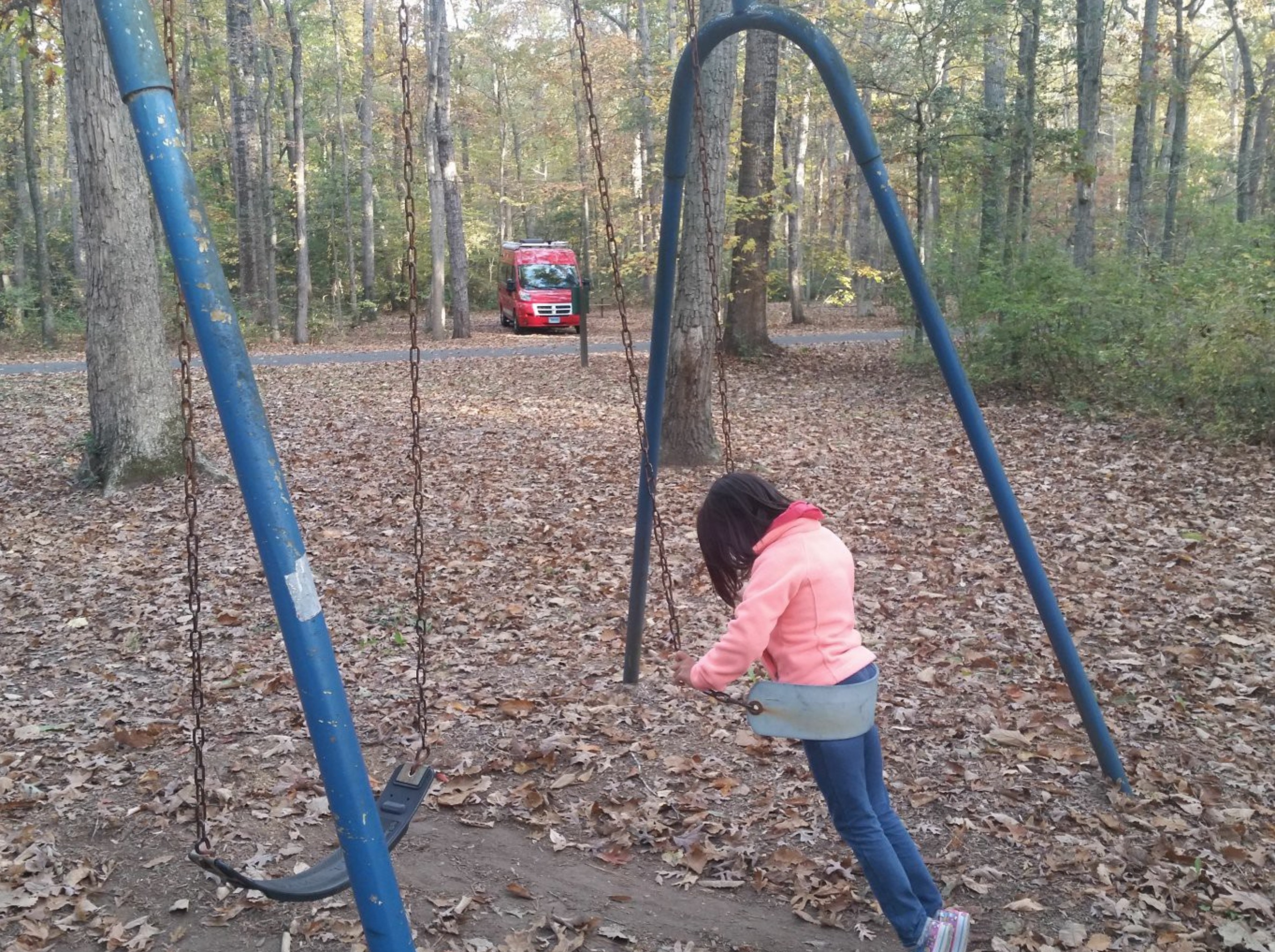 Girl on a swing set with red Winnebago Travato parked in the background.