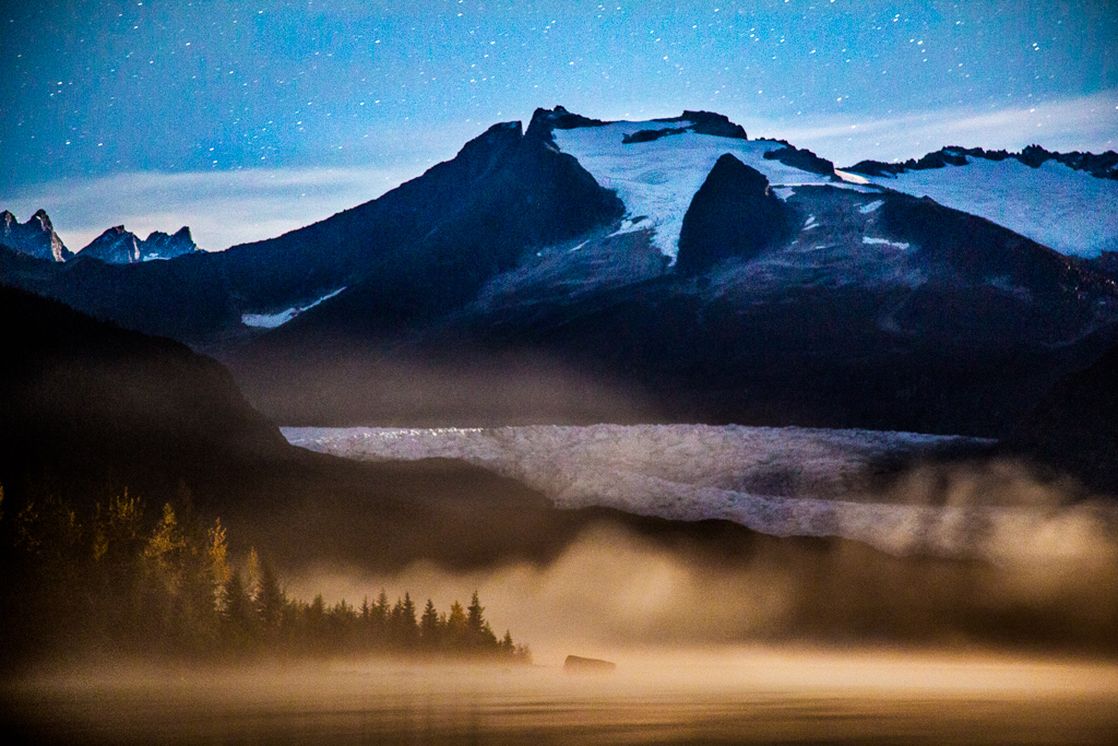 Starry night sky above, mountains and glacier meeting the fog covered water.