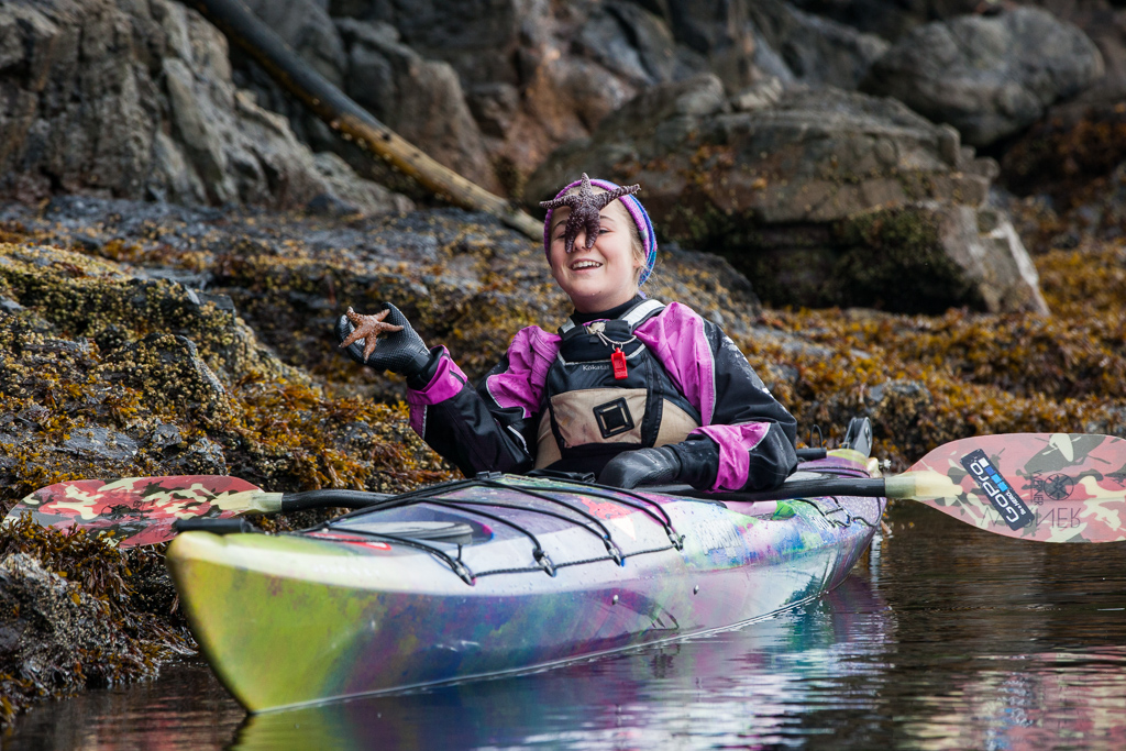 Abby on the water in her kayak along a rock holding one starfish, and with another one resting on her face.