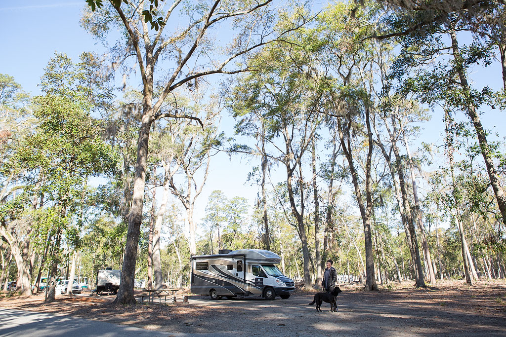 Woman and dog outside Winnebago View in tree filled campsite.