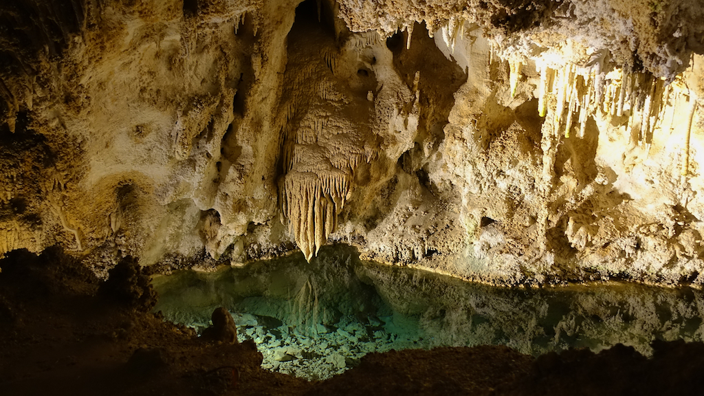 Incredible formations of stalagmites and stalactites and a crystal clear pool of water within the cavern lit up by lights.