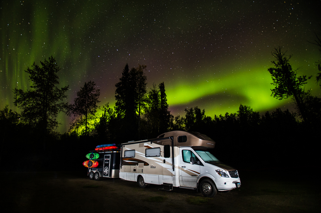 Winnebago View with trailer attached parked with the Northern Lights and star filled sky above.