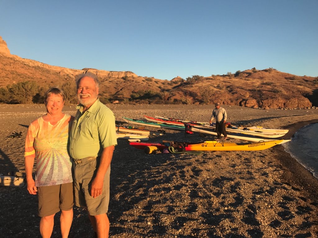 Couple smiling on the beach with paddle boards and kayaks behind them.