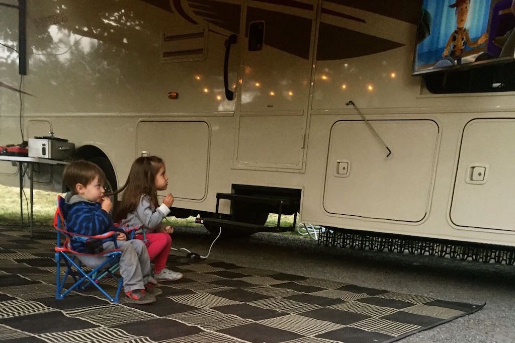 Two kids sitting in small camping chairs watching a movie on the exterior TV of the Winnebago Vista.