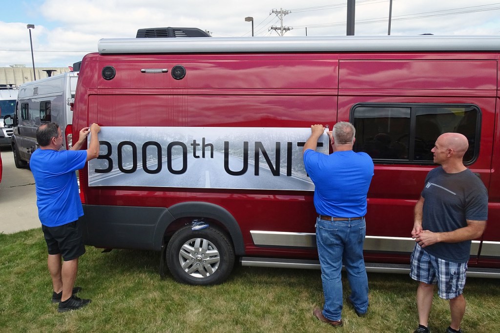 Two men hanging a sticker on a Winnebago class be van that reads "3,000th" unit.