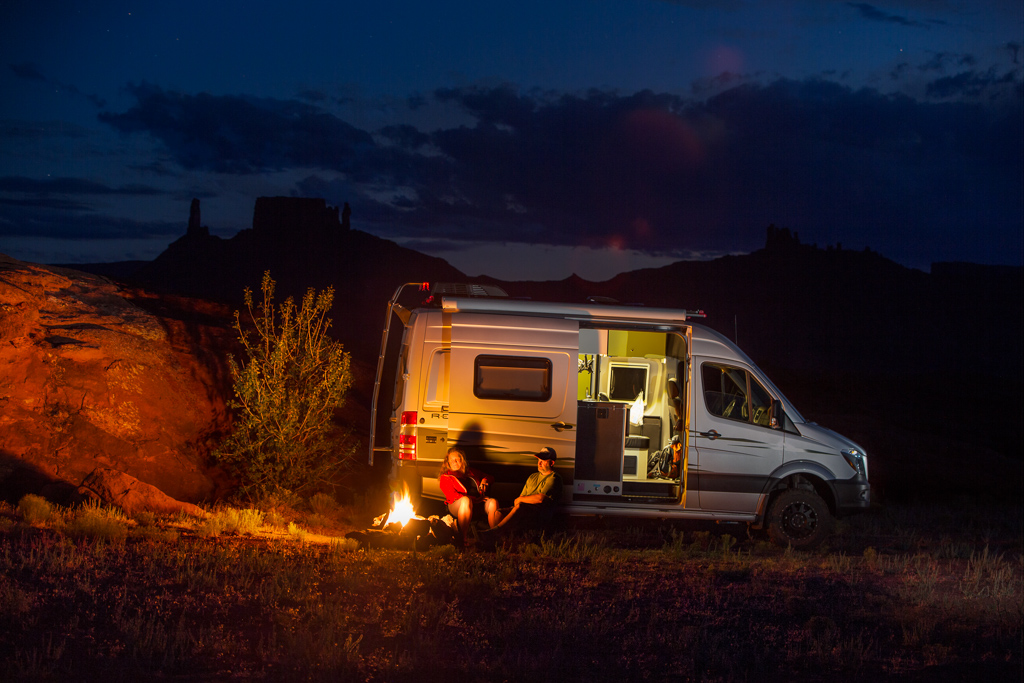 Couple sitting by a campfire outside their Winnebago Revel at night.