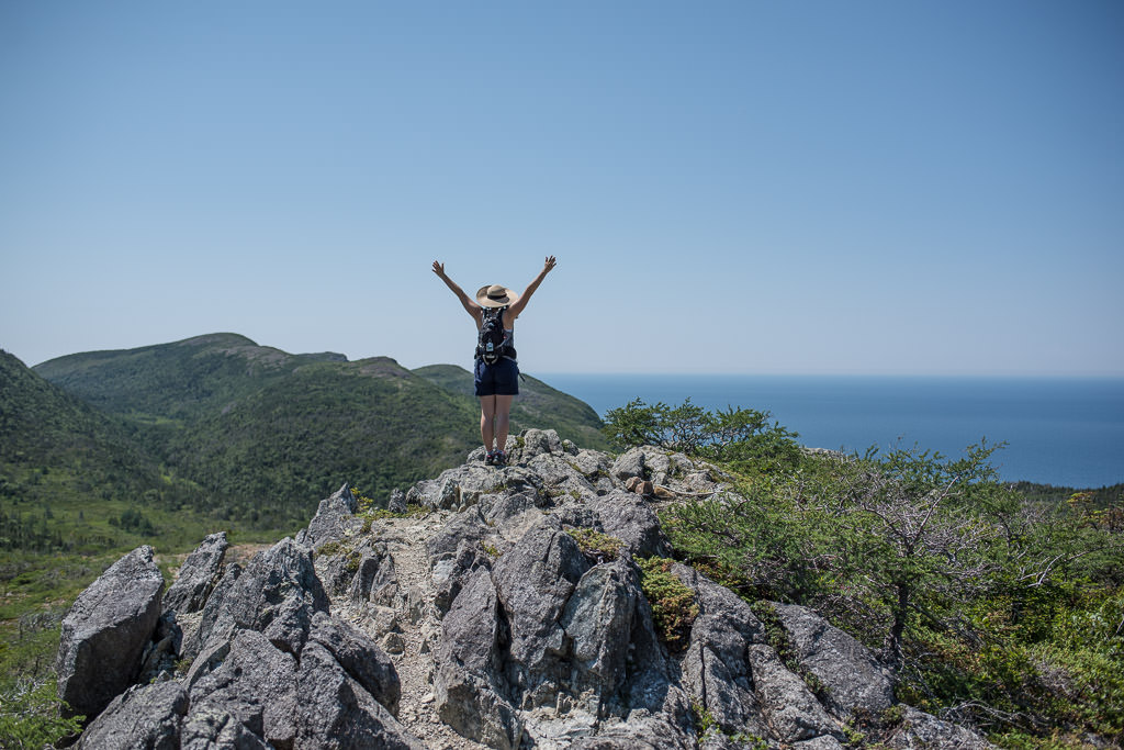 Woman standing with arms raised in the air atop a rocky hill with green hills and water