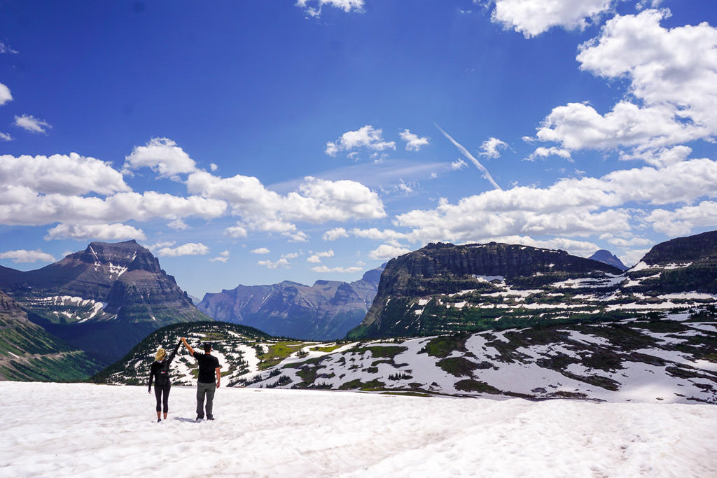 Couple holding hands in the air while standing on snow-covered ground and mountains ahead.
