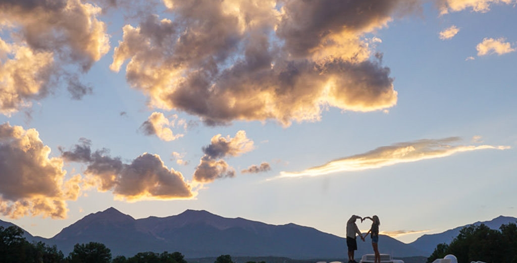 Couple forming a heart with their arms with background of mountains and beautiful clouds in the sky.