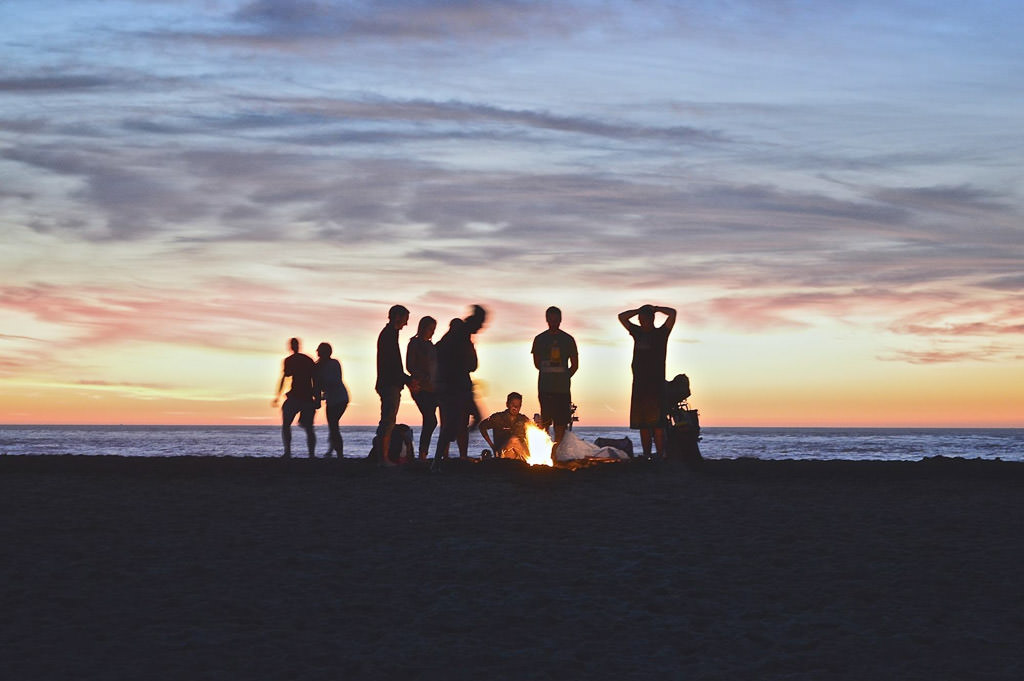 Group of people gathered around the fire on the beach at sunset.