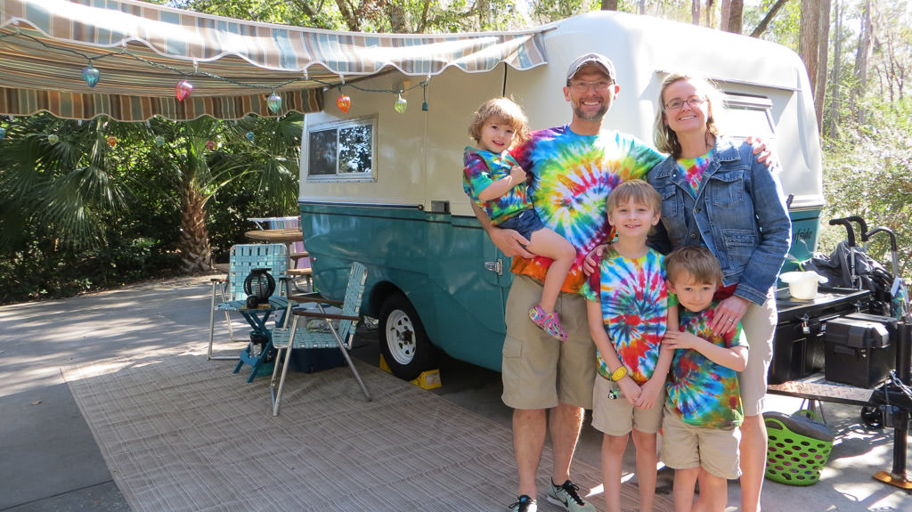 Family of five smiling for photo in front of travel trailer. Everyone is wearing tie dye shirts