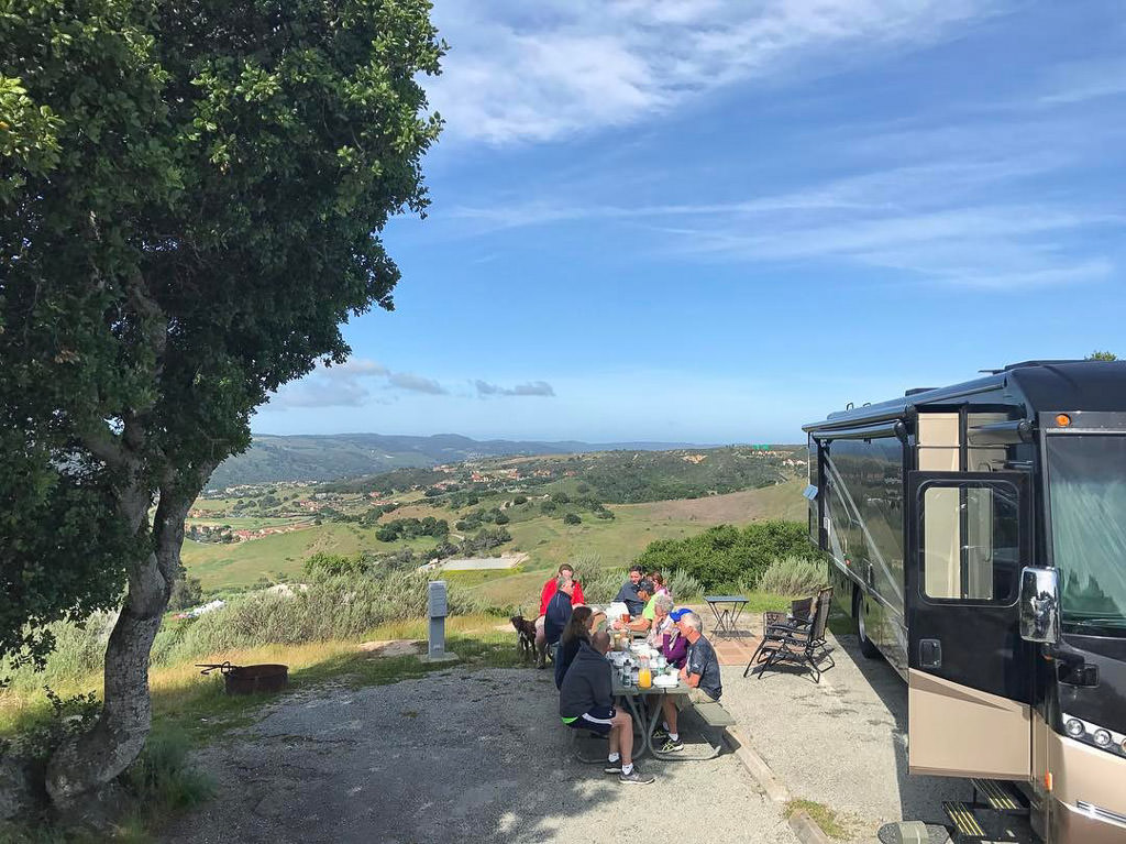 People gathered at a picnic table outside a motorhome with town and rolling hills in the background