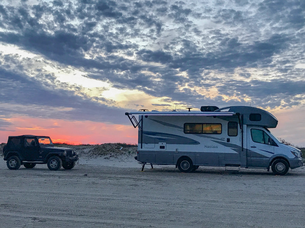 Winnebago Navion and vehicle parked on the sand with colorful sky in the background