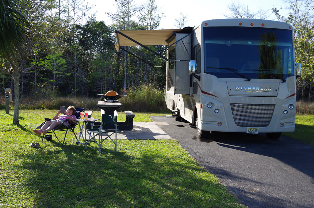 Winnebago motorhome parked on paved pad in Midway Campground with woman outside enjoying the sun and a book.
