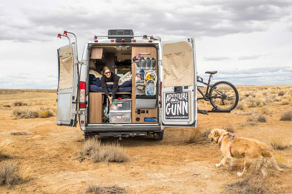 Van with backdoors open and woman leaning out the back interacting with their dog in a wide open desert area