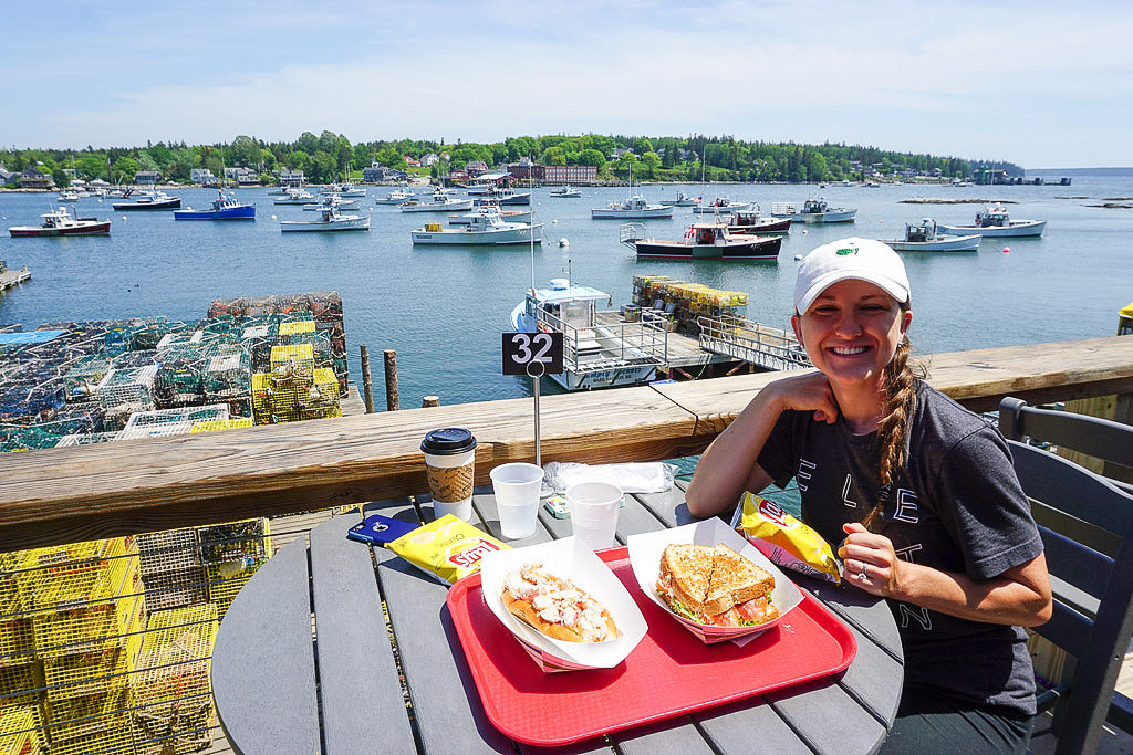 Woman eating meal at a restaurant on the water with boats on the harbor in the background.
