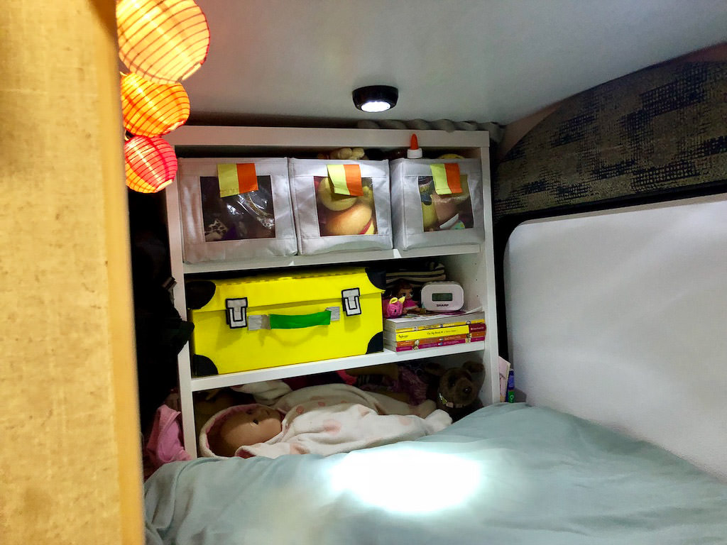 Storage for toys at the end of the bed. Three bins on the top shelf, a yellow suitcase on the second, and a bed for dolls on the third.
