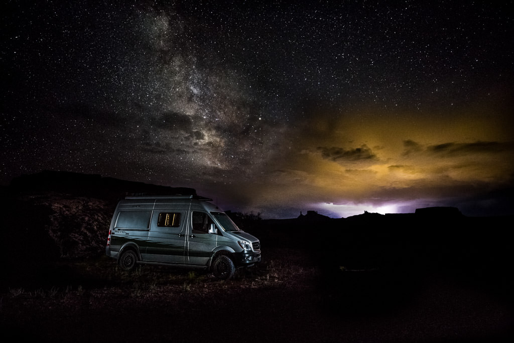 Winnebago Revel parked beneath the night sky with the Milky Way and an approaching storm lighting the sky.