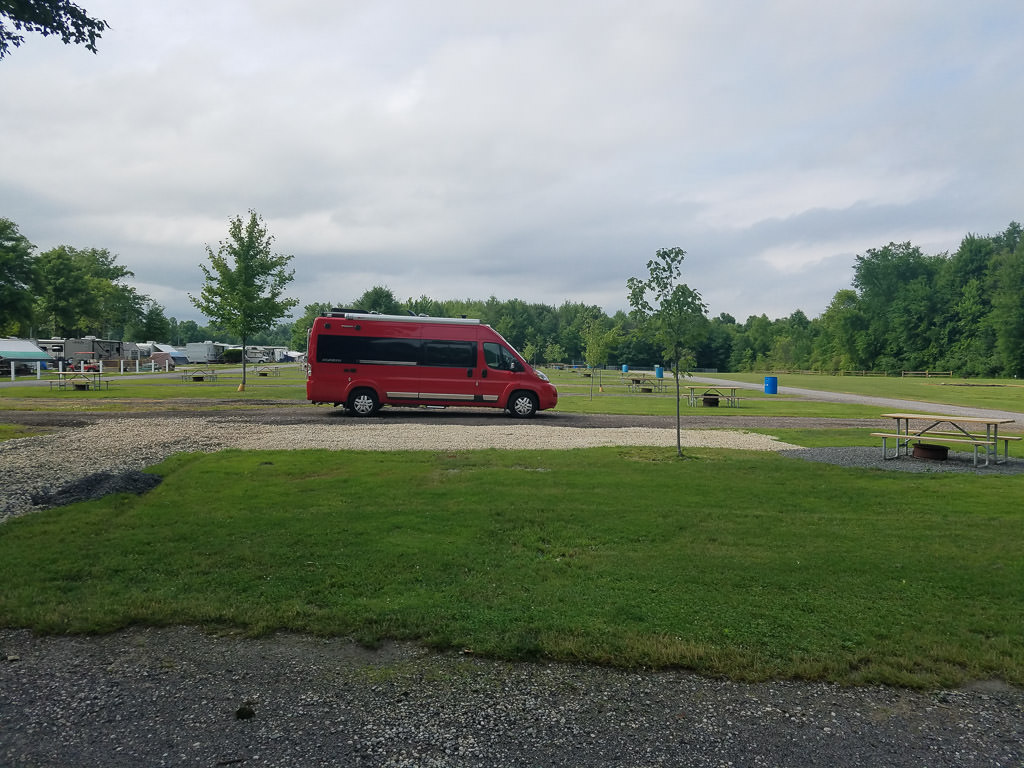 Red Winnebago Travato parked in middle of grassy campground