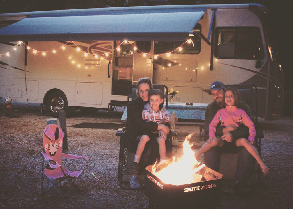 Winnebago Vista with lights strung along the awning and family sitting outside by a campfire.