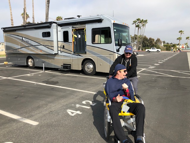 Mike pushing Patrick in his wheelchair with parked Accessibility Enhanced Winnebago Forza parked in parking lot behind them..