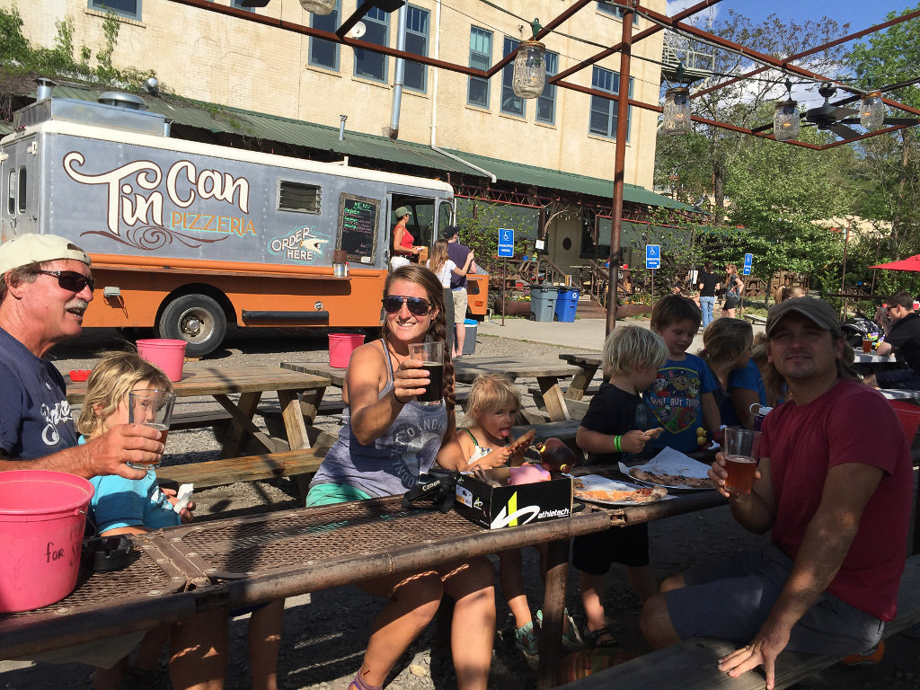 People gathered at a picnic table with food and drinks in front of the Tin Can Pizzeria food truck.