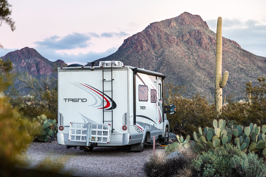 Winnebago Trend parked at campsite next to cacti with mountains ahead.