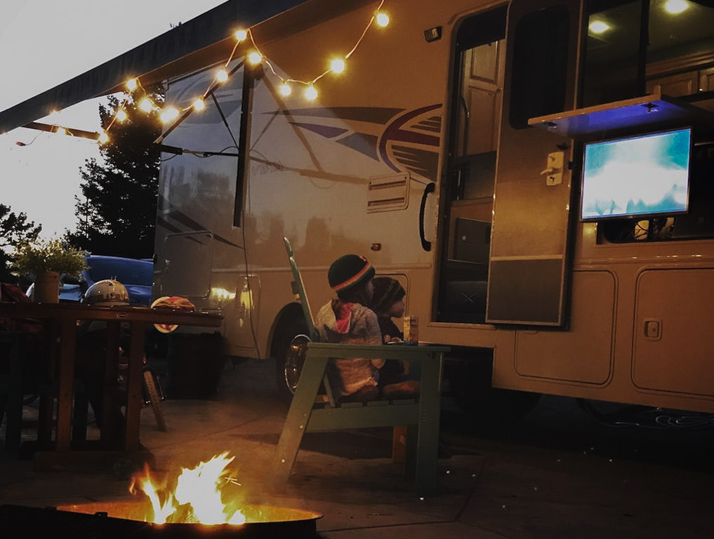 Two kids sitting in chairs outside Winnebago Vista with campfire burning and outside TV on.