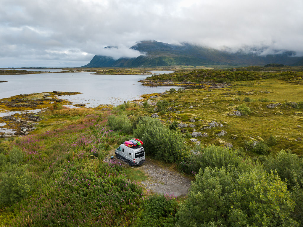 Winnebago Revel parked alone in grassy area near water with mountains in view.