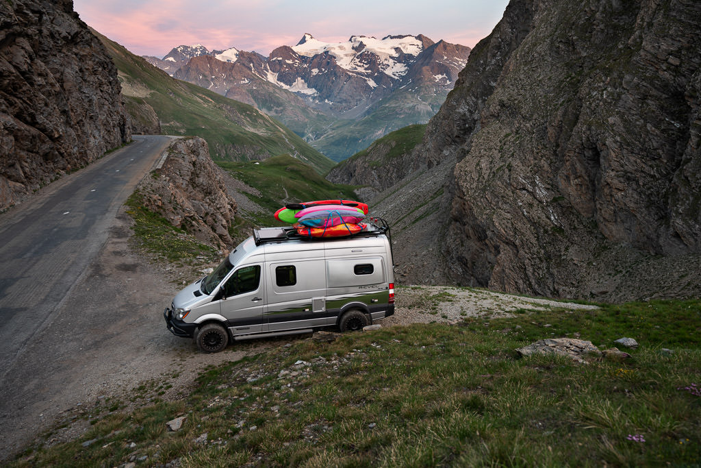Winnebago Revel parked to side of narrow road with snow capped mountains in the background.