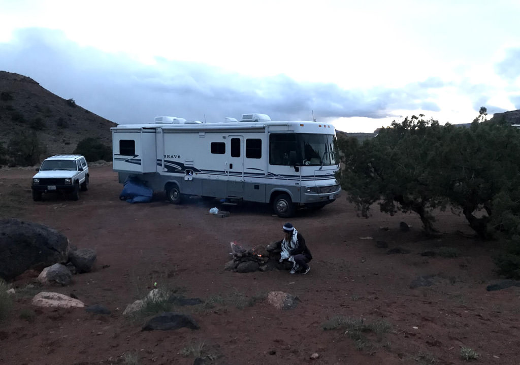 Winnebago Brave parked in rough desert looking landscape with woman kneeling next to a small fire.